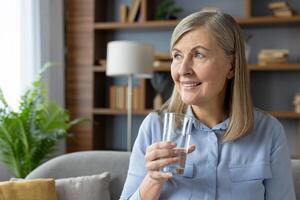 A mature woman with grey hair smiles warmly while holding a glass of water, seated comfortably in a well-lit living room decorated with plants and books. photo