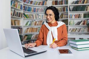 inspirado hembra estudiante sentado en biblioteca por escritorio con ordenador portátil y libros mientras haciendo notas por mano en papel hoja. interesado dama acecho en línea seminario web conferencia y haciendo observaciones para memorización foto