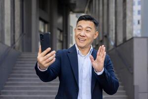 alegre asiático empresario atractivo en un llamada utilizando su teléfono inteligente, sonriente y ondulación Hola al aire libre. foto