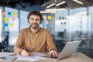 A confident male professional smiles while sitting at his desk in a bright, contemporary office space filled with natural light and colorful notes. photo