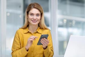 de cerca retrato de un joven mujer, oficina obrero, mujer de negocios sentado en el oficina a el mesa y utilizando el teléfono, sonriente a el cámara. foto