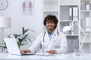 Portrait of young Indian doctor, man in white medical coat smiling and looking at camera, doctor sitting at table inside medical office of clinic, working with laptop. photo