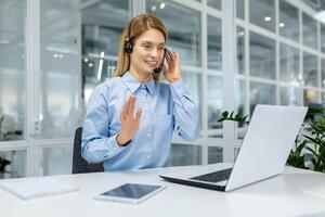 Friendly hr manager in headphones raising hand in greeting gesture while smiling to computer screen. Optimistic lady in light blue blouse welcoming remote companion and creating cozy atmosphere. photo