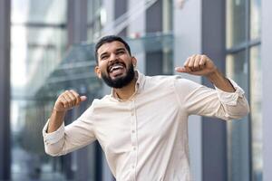 Close-up portrait of a young happy Muslim man standing outside on the street and rejoicing in good news and success, celebrating with joy and raising his hands up photo