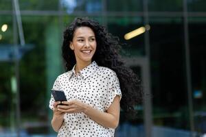 joven hermosa latín americano mujer caminando en el ciudad con teléfono en manos, negocio mujer después trabajando día, sonriente con satisfacción, estudiante utilizando solicitud en teléfono inteligente foto