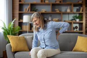 Mature lady feeling spinal muscles pain attack and holding back while squinting eyes on sofa with cushions. Sick woman having problem with standing up and experiencing kidney ache in living room. photo