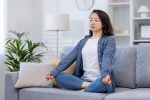 joven hermosa asiático mujer solo a hogar, meditando sentado en sofá en vivo habitación en loto posición, sentado con ojos cerrado a hogar. foto