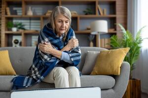 Sick senior lady hugging herself while sitting covered with checkered blanket next to electronic radiator. Worried female trying getting warm while waiting for beginning of central heating season. photo