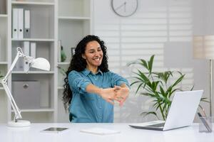 alegre mujer en un azul camisa tomando un extensión descanso a su limpio, organizado espacio de trabajo con ordenador portátil y oficina suministros. foto