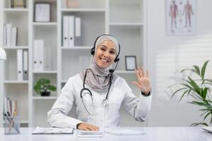 A cheerful female doctor in a white coat and hijab greets warmly during a call from her modern office. Her friendly demeanor and professional setting inspire trust and comfort. photo