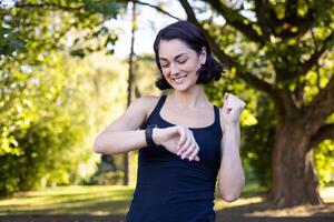 Joyful female runner checking her smartwatch, monitoring performance after training in a sunlit park. photo