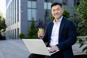 Professional Asian businessman working on laptop outside modern office building with coffee in hand, displaying success and mobility. photo