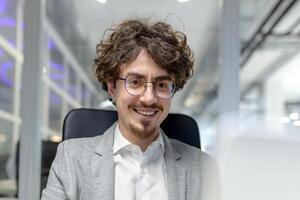 Close-up portrait of a smiling young businessman, developer man in a suit sitting in a modern office at a desk and working on a laptop. photo