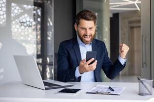 Successful and happy young businessman sitting in modern office at desk, looking at mobile phone and happy with achievement, showing victory gesture with hand. photo