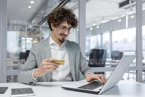 Confident young male entrepreneur with curly hair engaged in online financial operations, using a credit card and laptop at his workplace. photo