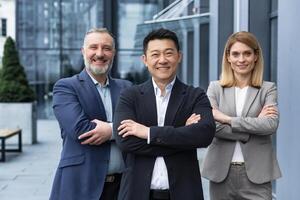 Successful diverse business team, three workers smiling and looking at camera, dream team with asian boss outside office building, colleagues in business suits photo