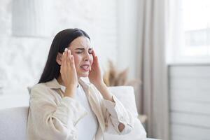 A woman suffers from a severe headache,pressure. She is sitting at home, holding her head with hands photo