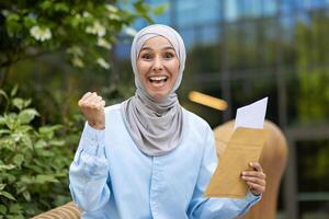 Vibrant image of a joyful Muslim woman wearing a hijab, exuberantly celebrating while holding an envelope outdoors with a blurred green background. photo