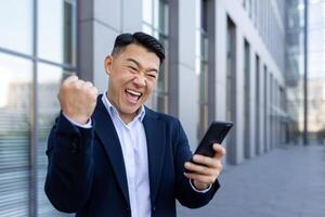 de cerca retrato de un asiático joven negocio hombre en pie en un traje cerca un oficina edificio, participación un teléfono en su manos y alegría en éxito, demostración un victoria gesto con su mano. foto