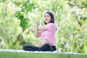 asiático embarazada mujer meditando mientras sentado en el parque, mujer haciendo extensión ejercicios al aire libre, meditando en maternidad foto