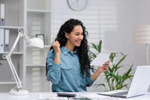 emocionado Hispano mujer de negocios celebrando un exitoso momento mientras trabajando desde su hogar oficina, expresando alegría y éxito. foto