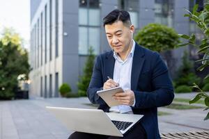 Asian young businessman working outside office center, sitting on bench wearing headphones, holding laptop and making notes in notebook. photo