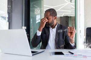 sobrecargado de trabajo hombre a lugar de trabajo dentro oficina, africano americano empresario trabajando sentado con ordenador portátil a trabajar, jefe tomando apagado lentes y masajear ojos. foto