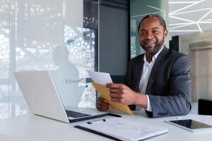 retrato de exitoso africano americano empresario financiero a lugar de trabajo dentro oficina edificio, masculino maduro adulto sonriente y mirando a cámara, jefe recibido correo sobre mensaje. foto