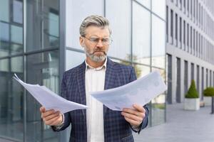 Angry senior boss standing on the street in front of the office, holding work documents, problems at work, bad news. photo