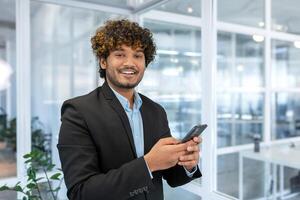 retrato de exitoso Hispano empresario inversor, hombre sonriente y mirando a cámara, jefe en traje a trabajo dentro oficina participación teléfono inteligente foto