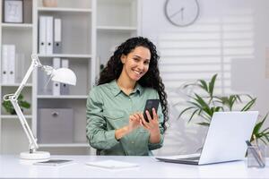 profesional Hispano mujer trabajando en un brillante oficina espacio con un ordenador portátil y móvil teléfono, mostrando eficiencia y multitarea habilidades. foto