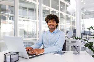 Portrait of young programmer inside office with laptop, indian man smiling and looking at camera, man typing on keyboard, writing code for software, happy with achievement results. photo