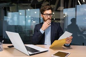 Sad and upset mature businessman received an envelope by mail with a bad news notification letter, boss in business suit working inside office using laptop wearing glasses and beard. photo