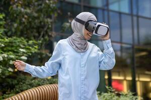 sonriente musulmán dama en hijab utilizando virtual realidad auriculares afuera, experimentando moderno tecnología en un natural ajuste. foto
