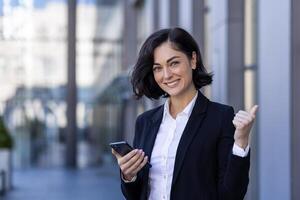 Portrait of a young successful business woman standing near an office center, holding a phone, showing a super finger and smiling at the camera. photo
