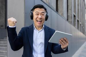 retrato de un contento asiático hombre en un negocio traje y auriculares, en pie fuera de un edificio, participación un tableta en su mano y diciendo éxito a el cámara. foto