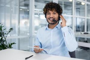 un alegre masculino cliente Servicio representante con Rizado pelo vistiendo un auriculares, sonriente y trabajando en un moderno oficina ambiente. foto