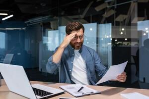 Upset businessman behind paper work inside modern office, mature man with beard reading financial reports and account documents unhappy with results and disappointed with achievements. photo