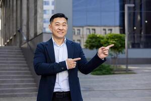 Smiling Asian businessman in a suit pointing with both hands to the side, standing on a city street with buildings in the background. photo