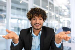 A successful hispanic man inside the office is looking at the web camera, talking on a call with colleagues, gesturing and smiling an online meeting with partners, talking to customers, pov photo