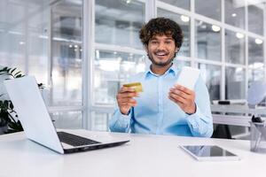 contento joven hombre sentado en oficina a escritorio con teléfono y crédito tarjeta. hace en línea compras, paga facturas. sonriente mirando a el cámara. foto