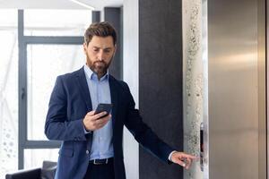 Focused businessman in a suit using smartphone while waiting for an elevator in a modern office building interior. photo