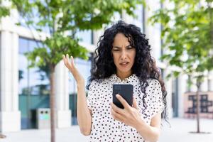 Sad disappointed woman received online notification with bad news on her phone, businesswoman walking outside office building, using application on smartphone, reading social media unsatisfied. photo