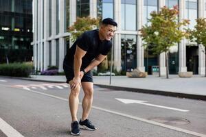 joven asiático masculino corredor en pie en el medio de el calle doblado encima, participación su pierna y rodilla, tirado un músculo, ocultación y sufrimiento desde dolor. foto