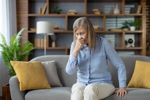 Caucasian woman with low immunity getting seasonal cold and sneezing in paper tissue at home. Mature lady trying to deal with influenza and having running nose while sitting with closed eyes. photo