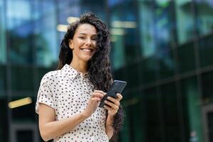 Contemporary young adult female engrossed in smartphone usage against the backdrop of a modern glass building. photo