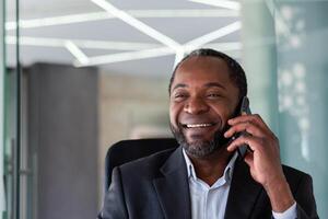 maduro experimentado exitoso jefe hablando en el teléfono cerca arriba, africano americano hombre sonriente mirando a ventana, trabajando a lugar de trabajo dentro oficina con ordenador portátil en negocio traje. foto
