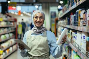 retrato de un hembra vendedora en un hiyab, un vendedor en un casa productos quimicos Departamento es sonriente y mirando a el cámara, participación un ordenador portátil tableta computadora en su manos foto