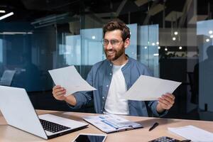 satisfecho con el resultado financiero empresario sostiene en su manos y examina contratos y informes sonriente y feliz, hombre en un casual camisa trabajos dentro el oficina utilizando un ordenador portátil en papeleo. foto