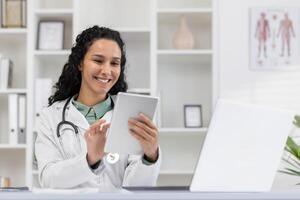 alegre hembra médico en un blanco Saco delicias en utilizando un digital tableta en su brillante médico oficina, ejemplificando profesional felicidad y digital cuidado de la salud. foto